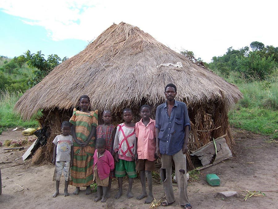 a family with a man, woman, and 5 children standing in front of a straw hut in a developing country